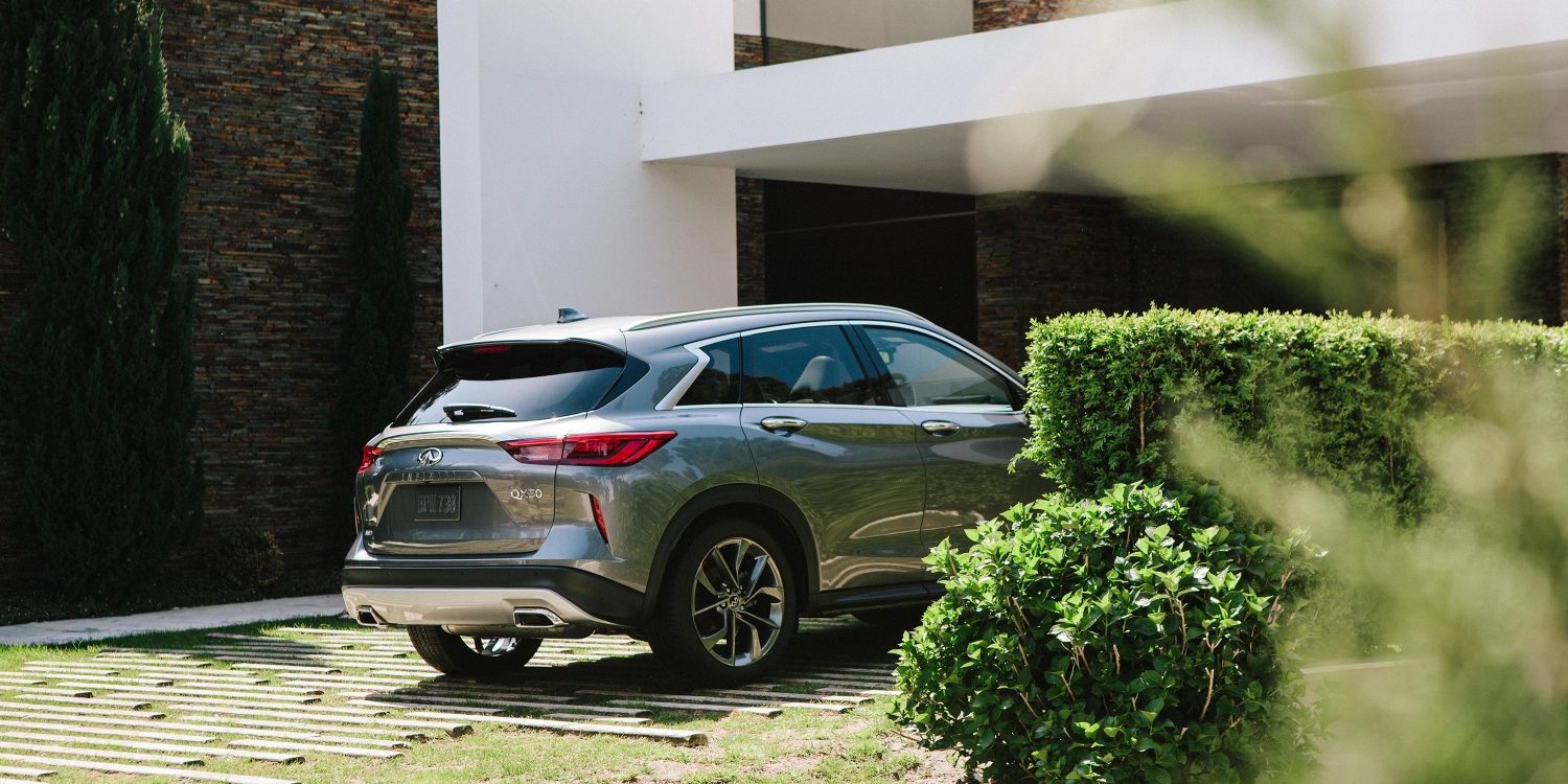 A gray INFINITI QX50 parked on a driveway in front of a home for sale