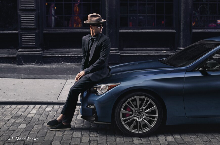 Man looking back and sitting on the hood of a luxurious 2022 INFINITI Q50 luxury sedan. U.S. model shown, Model may not be exactly as shown.