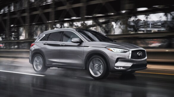 Side profile of a Graphite Shadow 2022 INFINITI QX50 Crossover driving fast on a wet road