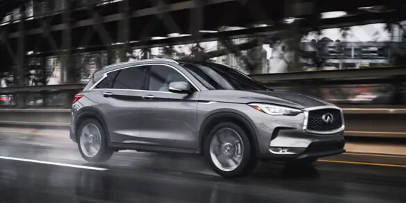 Side profile of a Graphite Shadow 2023 INFINITI QX50 Crossover driving fast on a wet road